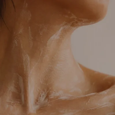A close-up shot of a person's neck and clavicle area with a white, foamy cleanser, soap, or exfoliating scrub applied to the skin. The product is lathered across the neck and upper chest.