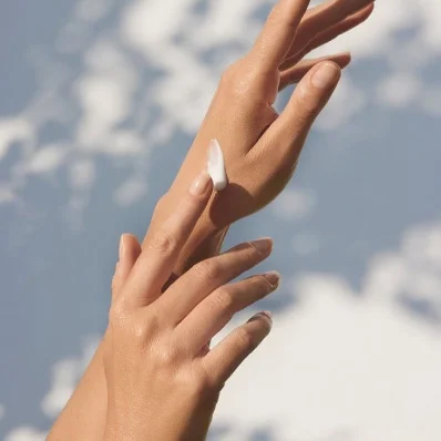A close-up shot of two hands applying a dollop of white cream or lotion. One hand is holding the other, and a finger is spreading the product on the back of the first hand. The background is a bright blue sky with soft white clouds, suggesting an outdoor or natural setting.