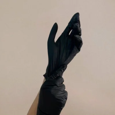 A close-up shot of a person's hand pulling on a black nitrile or latex medical glove. The hand is midway through pulling the glove up the wrist and is set against a plain, light beige or off-white background.