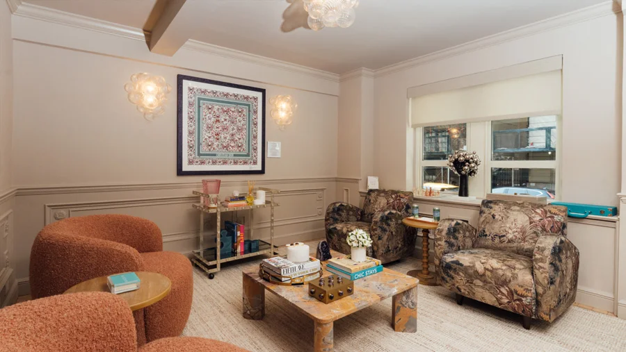 The elegant, warm waiting room of Dr. Kassir's office. The room features a cozy mix of seating, including two orange boucle armchairs and two floral patterned armchairs around a rustic coffee table. A large framed print hangs on the beige wall, flanked by bubble-style wall sconces. A glass and brass bar cart is near a chair.