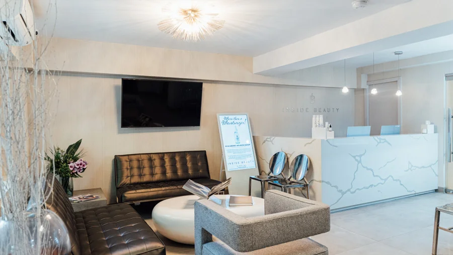 The bright, modern waiting area of a cosmetic or medical office called "INSIDE BEAUTY." The reception desk is white with gray veining. The waiting area has a black leather sofa, a gray armchair, a white coffee table, and a promotional poster. A large TV is on the wall, and the room is decorated with minimal, contemporary lighting.