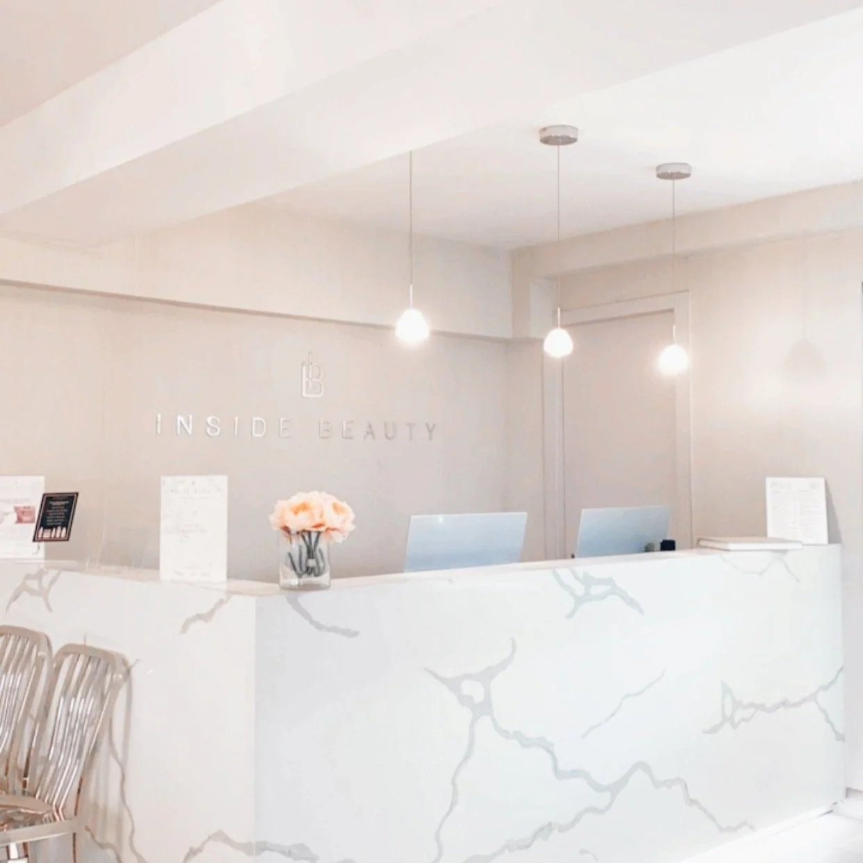 A bright, clean, and minimalist reception area for "INSIDE BEAUTY." The focal point is a large, white reception desk with gray marble-like veining. The logo and text "INSIDE BEAUTY" are mounted on the wall behind the desk. Two computers and a vase of light pink flowers sit on the countertop. Three simple pendant lights hang over the desk, and a few silver chairs are visible in the foreground.