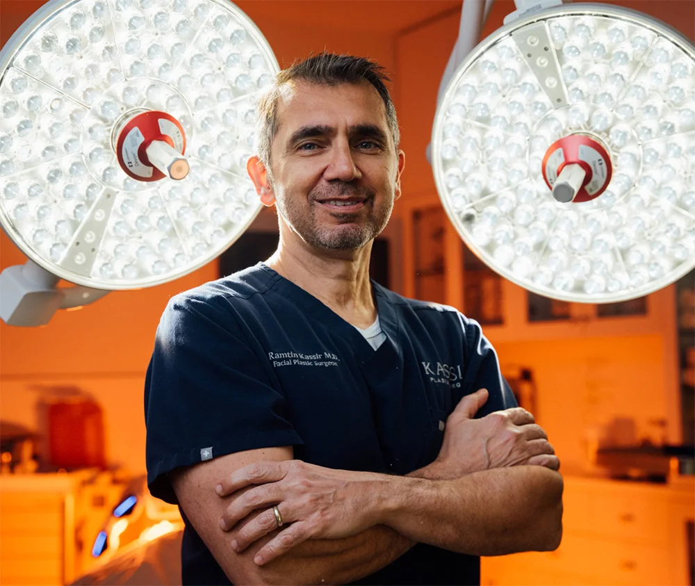 A portrait of Dr. Kassir, a facial plastic surgeon, standing in an operating room under two bright surgical lights. He is wearing dark blue scrubs with his arms crossed and smiling directly at the camera. His name and the Kassir Plastic Surgery logo are visible on his scrubs.