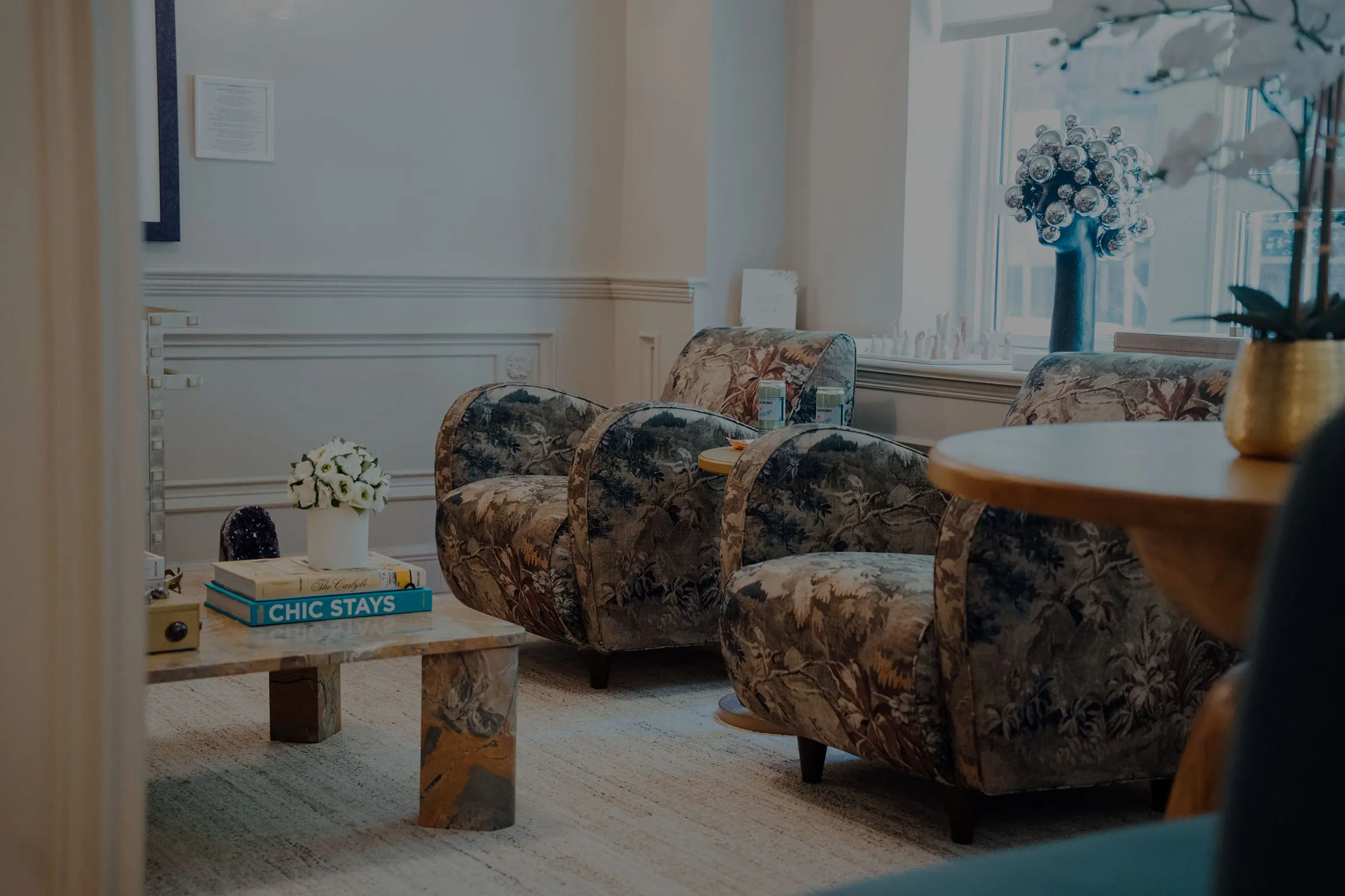 A bright, luxurious waiting room featuring two plush, upholstered floral-patterned armchairs. In the foreground, there is a marble coffee table with books and a small floral arrangement. Natural light comes through a large window on the right. - Kassir Wellness Center in New York City, NY