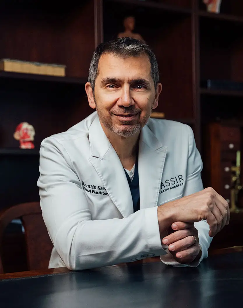 A formal portrait of Dr. Kassir seated at a dark desk, wearing a white lab coat with his arms crossed and hands clasped on the desk. He is looking directly at the camera and smiling slightly. The background is a dark wood shelving unit with a few decorative objects. - Kassir in New York City, NY