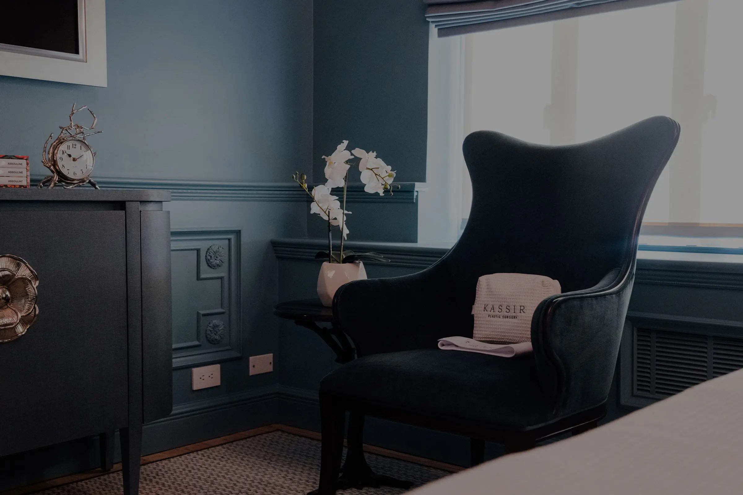 A close-up interior shot of a corner in a luxurious, teal-blue painted room. The image features a dark, velvet wingback chair with a white towel and a bag from "KASSIR" resting on it. Next to the chair is a small side table holding a white orchid plant. To the left is a dark blue credenza with a large brass flower-shaped pull and a vintage-style clock. Bright daylight streams in through the large window in the background.