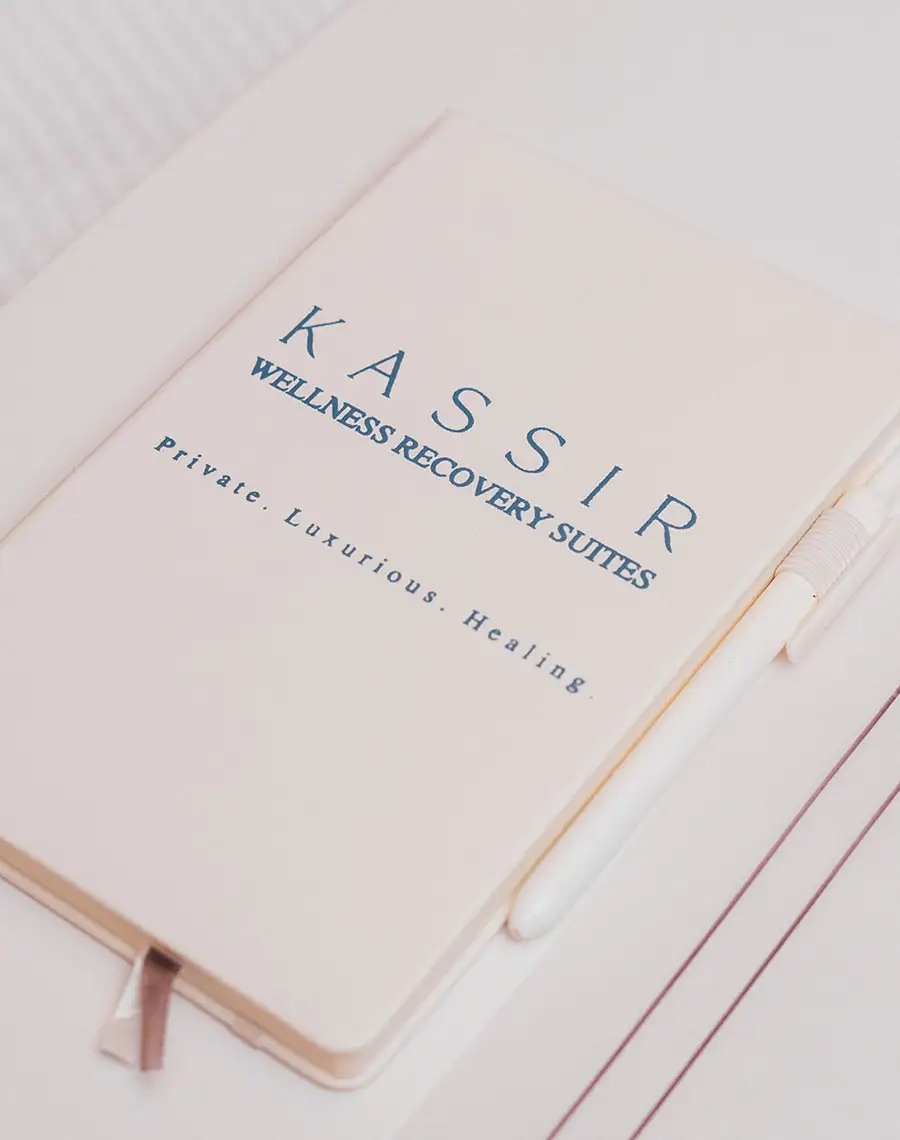 A close-up image of a creamy white notebook with a white pen resting on it. The text on the cover is in blue, reading "KASSIR WELLNESS RECOVERY SUITES" and, in smaller text below, "Private. Luxurious. Healing." - Wellness in New York City, NY