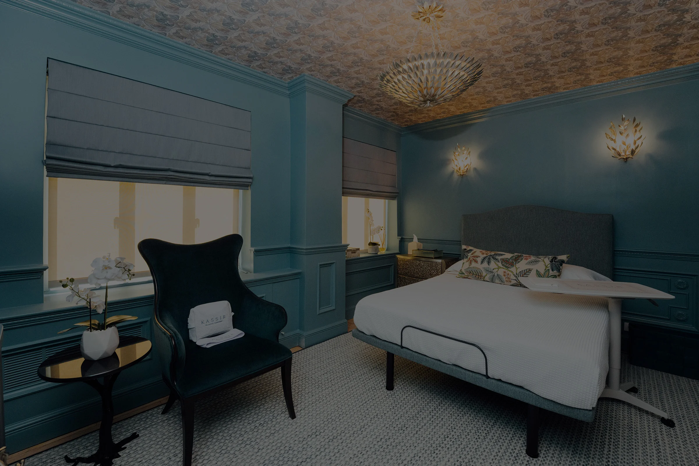 Elegant medical spa treatment room featuring deep teal paneled walls, a patterned metallic ceiling, and warm decorative lighting. A treatment bed with crisp white linens sits beside a dark upholstered chair and a small table with white orchids, creating a luxurious and calming wellness environment.